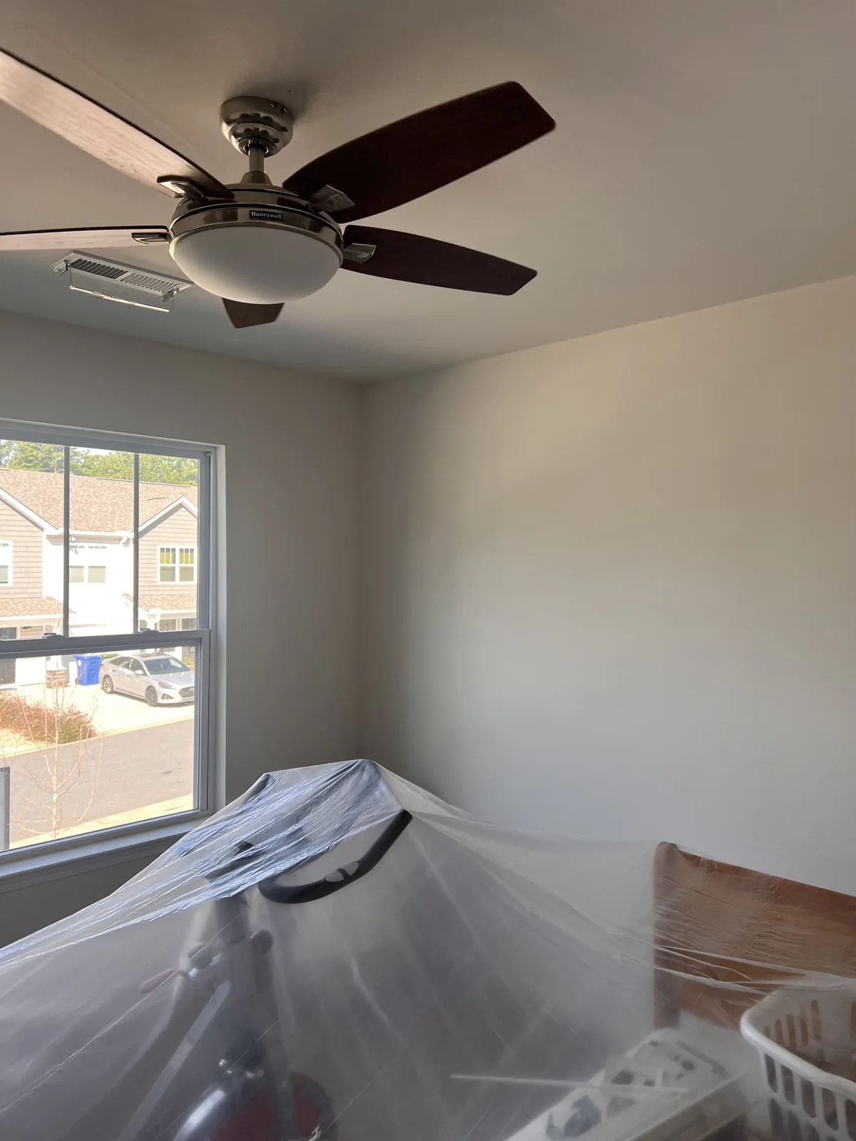 Light-colored wall beside a window, with a ceiling fan and furniture covered in protective plastic